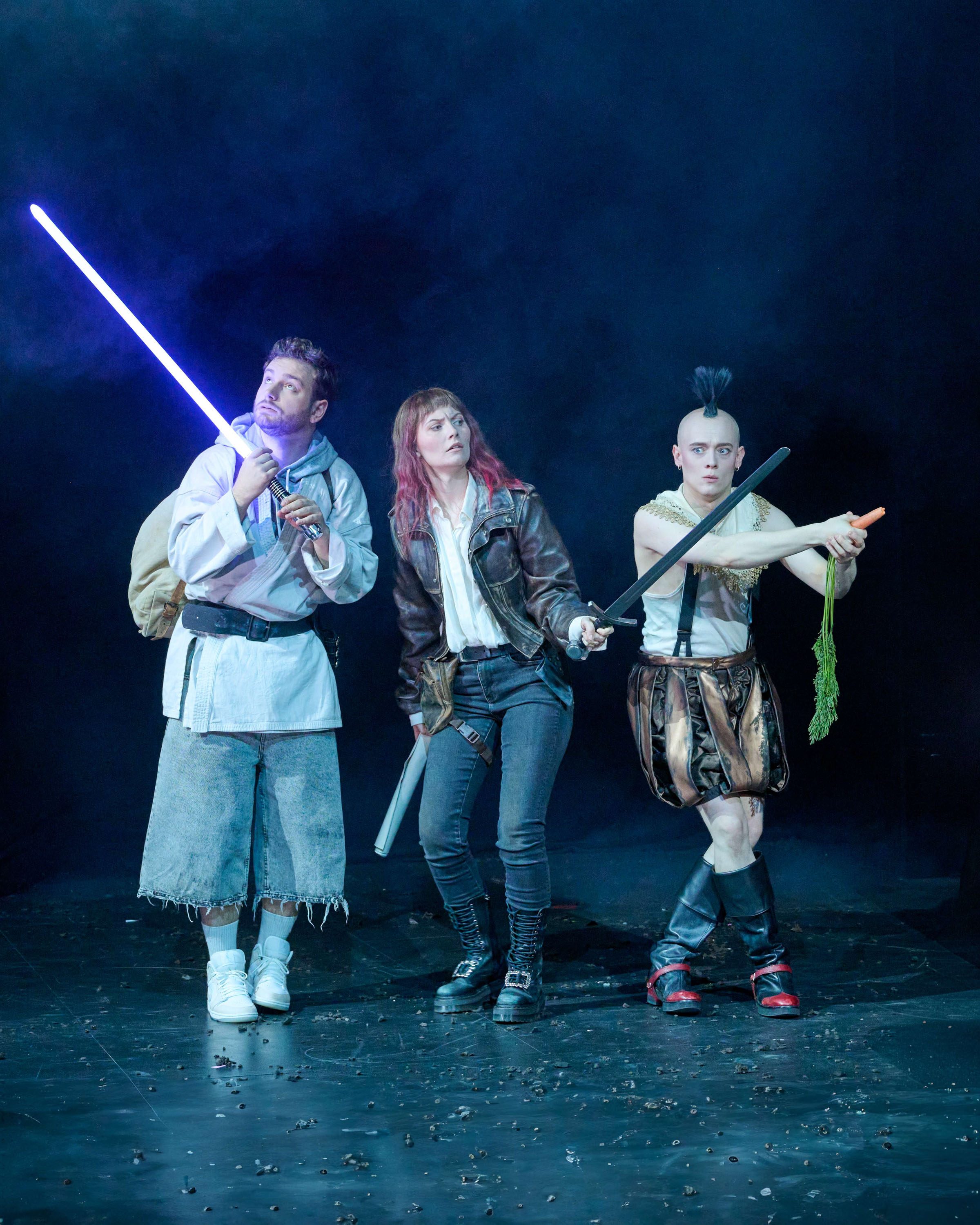 Three performers stand dramatically on stage. One holds a glowing purple sword, another has a sword in hand and wears a leather jacket, while the third brandishes a carrot and wears a unique costume. The background is dark with a smoky atmosphere, enhancing the theatrical scene.