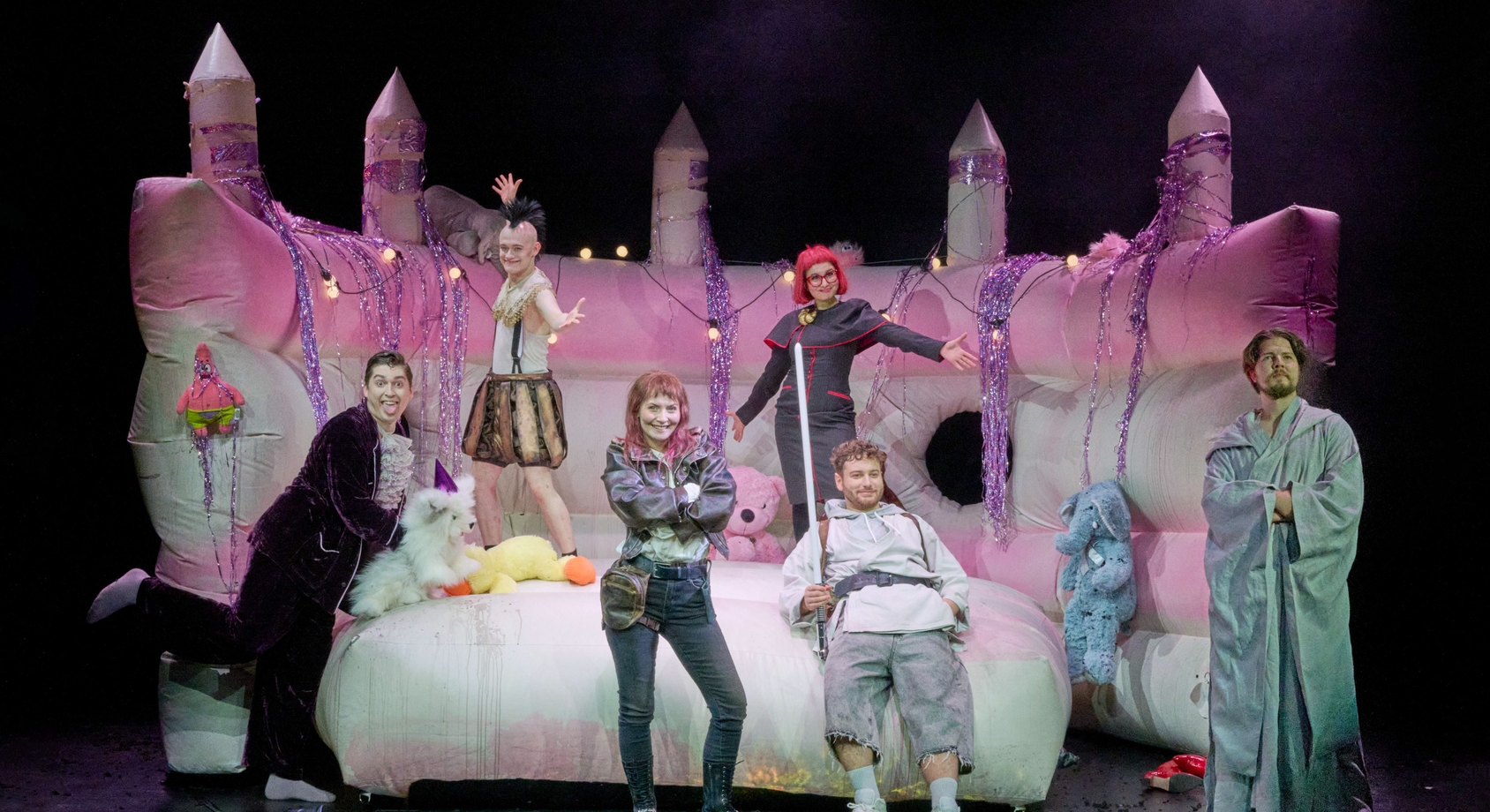 A group of six performers poses on a colorful inflatable castle adorned with lights and decorations. They exhibit playful expressions and unique costumes, engaging in a lively scene, with various plush toys scattered around them. The environment suggests a whimsical, festive atmosphere.